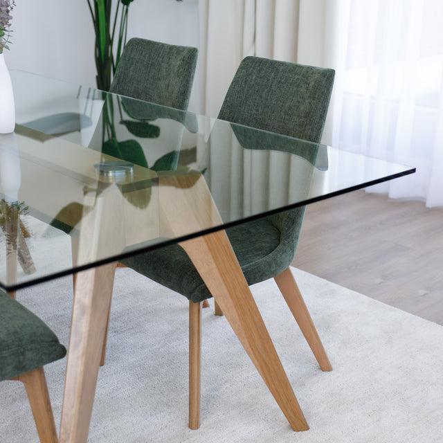 Table à manger en verre avec pieds en bois et chaises en tissu vert, style moderne dans un intérieur lumineux.