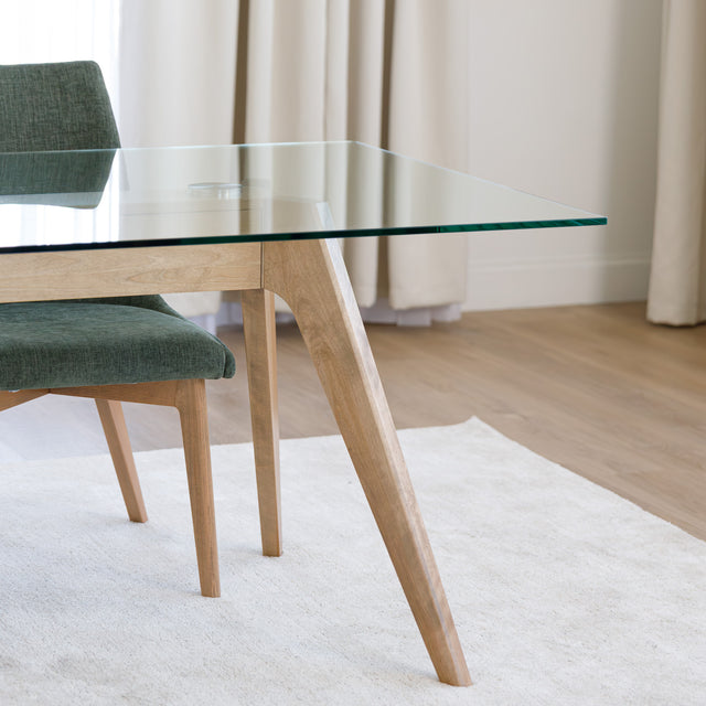 Table à manger en verre avec pieds en bois et chaises en tissu vert, style moderne dans un intérieur lumineux.
