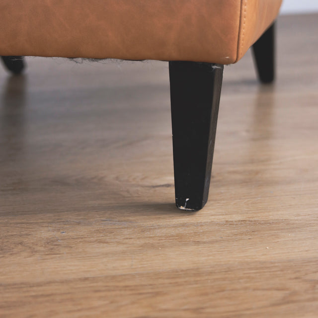 Image rapprochée d'une patte d'un fauteuil moderne en cuir végane cognac avec pieds noirs sur sol en bois.