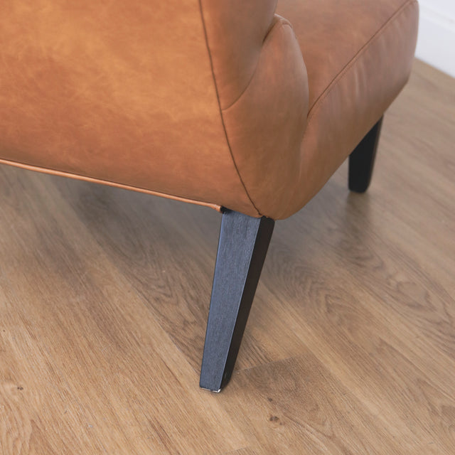 Image rapprochée d'une patte d'un fauteuil moderne en cuir végane cognac avec pieds noirs sur sol en bois.