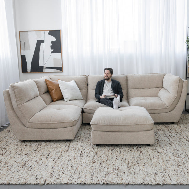 Canape sectionnel beige moderne avec pouf assorti et personne assise sur tapis texture clair.