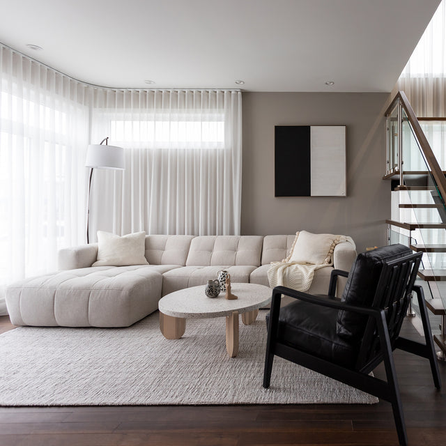 Salon moderne avec canape d angle beige clair matelasse, fauteuil noir et table ronde en bois clair.
