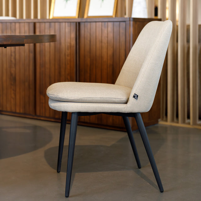 Chaise de salle à manger en tissu beige avec piètement noirs, placée près d’une table en bois dans un intérieur moderne.