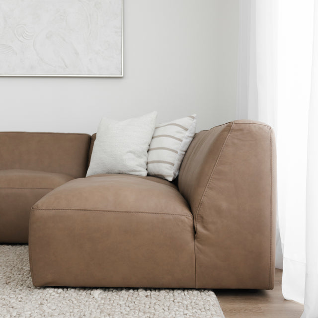 gros plan sur un canapé d’angle en cuir brun avec coussins beiges et rayés, posé sur un tapis épais texturé clair.
