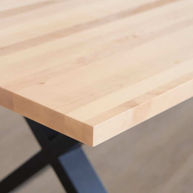 Image rapprochée d'une table rectangulaire en bois clair avec pieds en metal noir en X, design industriel elegant.