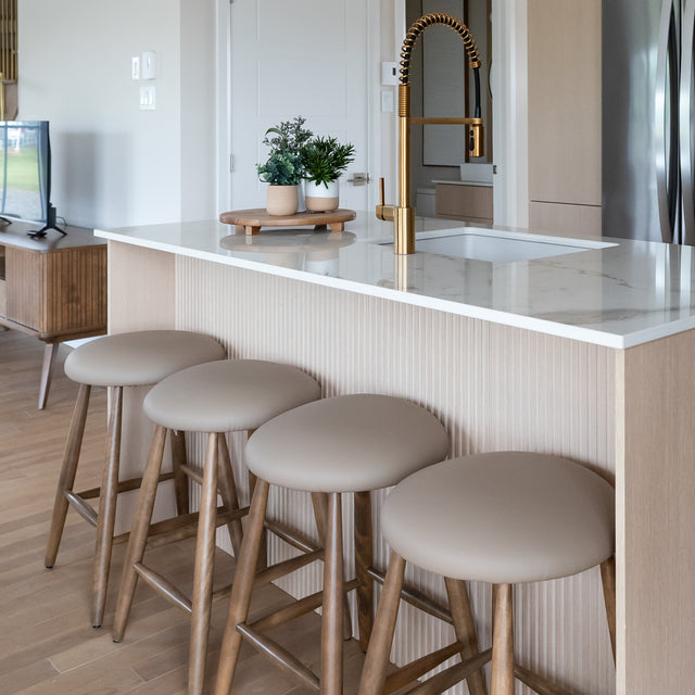 Quatre tabourets de bar en cuir végane taupe avec pieds en bois devant un ilot de cuisine moderne.