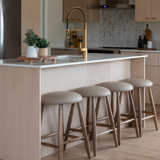 Quatre tabourets de bar en cuir végane taupe avec pieds en bois devant un ilot de cuisine moderne.