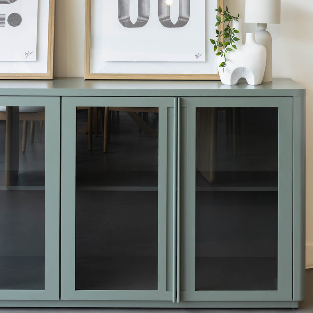 Buffet moderne vert sauge à portes vitrées fumées, décoré d’objets design dans un intérieur contemporain lumineux.