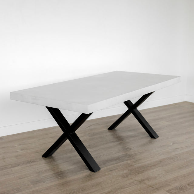 table rectangulaire moderne avec un plateau blanc mat épais et des pieds en métal noir en forme de X, posée sur un sol en bois clair devant un mur blanc.