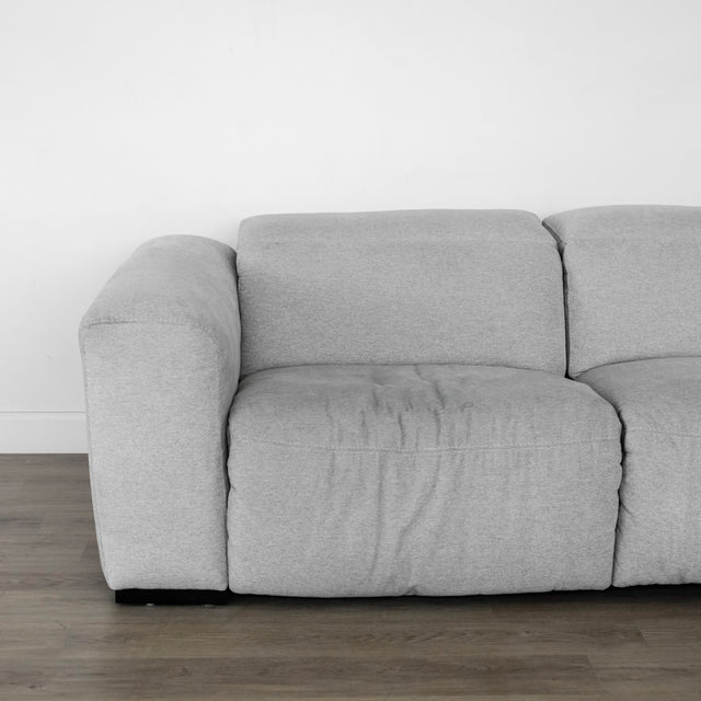 canapé en tissu gris clair au design contemporain, avec accoudoirs larges et coussins d’assise moelleux, posé sur un sol en bois clair devant un mur blanc.