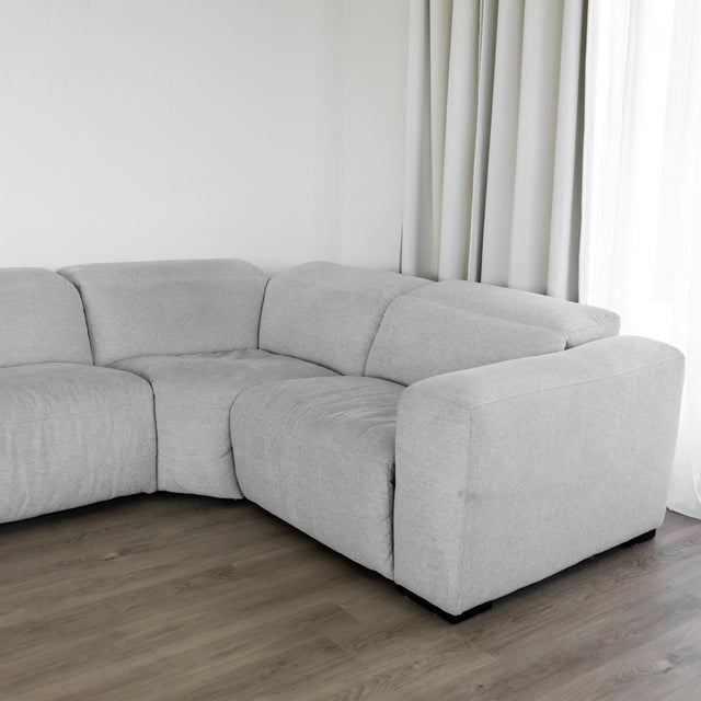 canapé d’angle moderne en tissu gris clair avec des formes arrondies et une assise profonde, placé sur un sol en bois clair devant un mur blanc et de longs rideaux beiges.