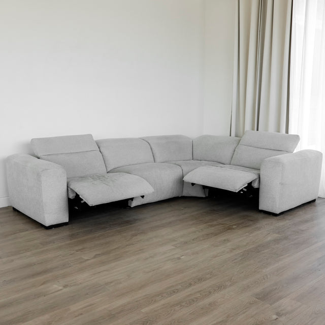 canapé d’angle en tissu gris clair avec deux assises inclinables dotées de repose-pieds relevés, design moderne et confort moelleux, placé sur un sol en bois clair près d’un mur blanc et de longs rideaux beiges.