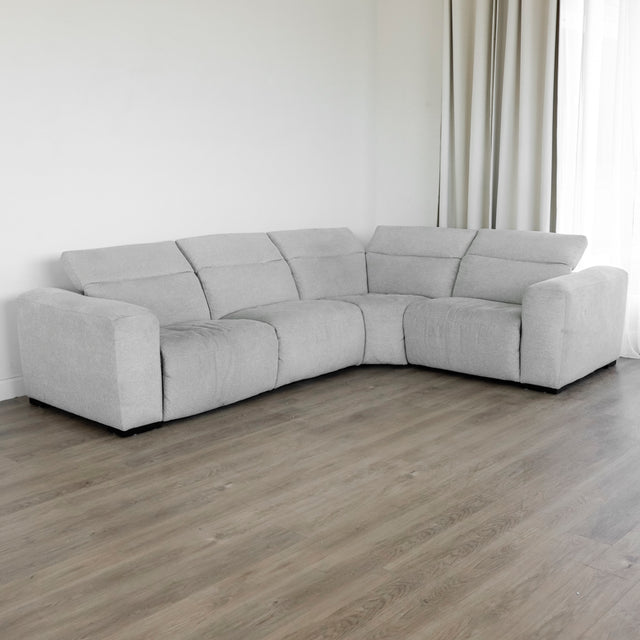 canapé d’angle en tissu gris clair au design contemporain, avec des accoudoirs larges, des dossiers inclinés et des coussins d’assise moelleux, placé sur un sol en bois clair devant un mur blanc et de longs rideaux beiges.