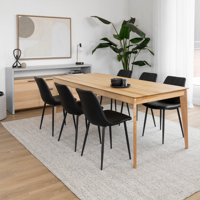 Table a manger en bois clair avec six chaises noires sur tapis beige dans salle moderne.