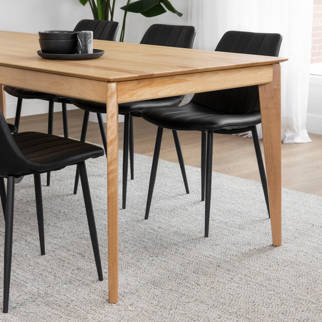 Table a manger en bois clair avec chaises noires sur tapis beige dans salle lumineuse.