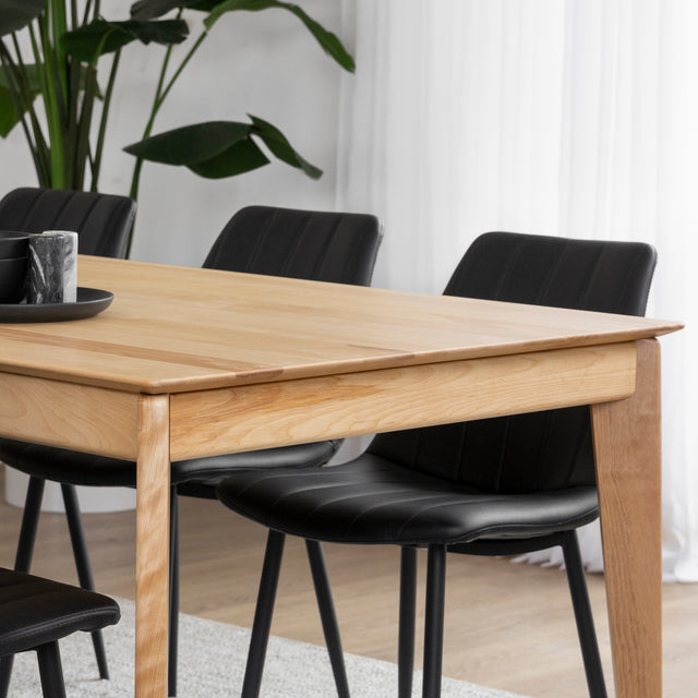 Table a manger en bois clair avec chaises noires sur tapis beige dans salle lumineuse.