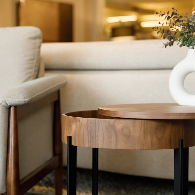 Table d’appoint ronde en bois noyer avec rangement, près d’un fauteuil beige.