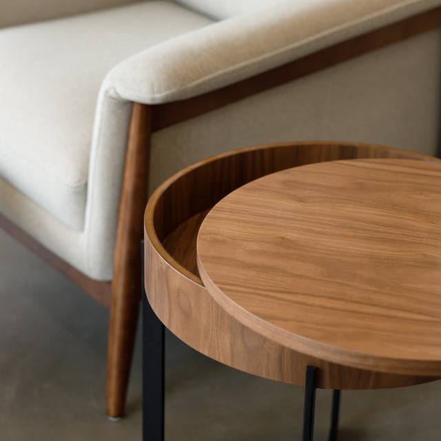 Table d’appoint ronde en bois noyer avec rangement, près d’un fauteuil beige.