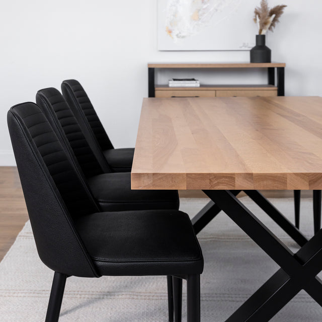 Table à manger en bois clair avec piètement en X métallique noir, entourée de six chaises noires dans une salle à manger moderne et épurée.