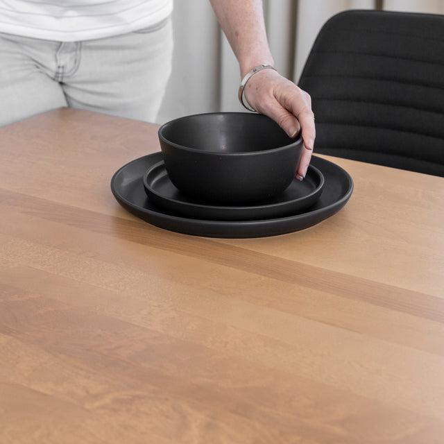 Gros plan sur une personne plaçant de la vaisselle noire mate sur une table en bois clair à la surface lisse et veinée.