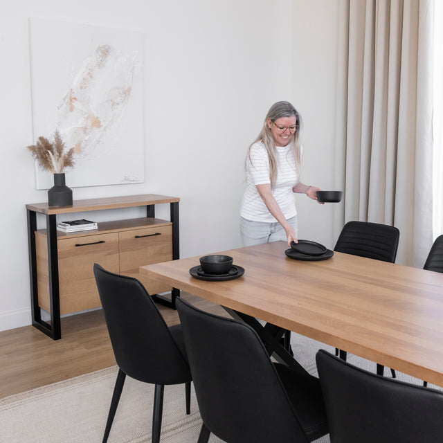 Femme dressant une table à manger en bois clair avec pieds métalliques noirs, entourée de chaises noires dans une salle à manger moderne et lumineuse.