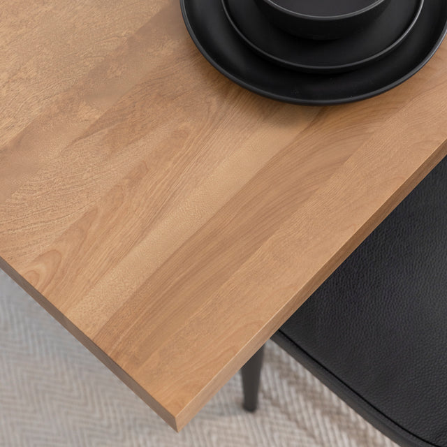 Gros plan sur un coin de table en bois clair à surface lisse et aux nuances naturelles, avec de la vaisselle noire mate et une chaise noire placée en dessous.