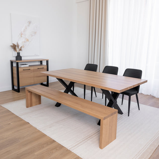 Deux personnes discutent à une table en bois clair avec pieds métalliques noirs en X, l’une assise sur une chaise noire et l’autre sur un banc assorti, dans une pièce lumineuse au style moderne.