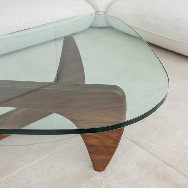 Table basse ovale avec plateau en verre et base en bois courbee devant canape clair.