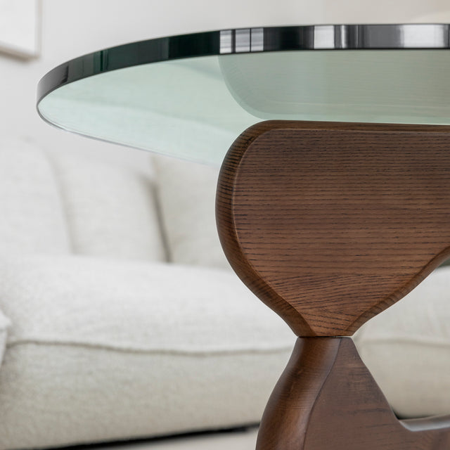 Table basse ovale avec plateau en verre et base en bois courbee devant canape clair.