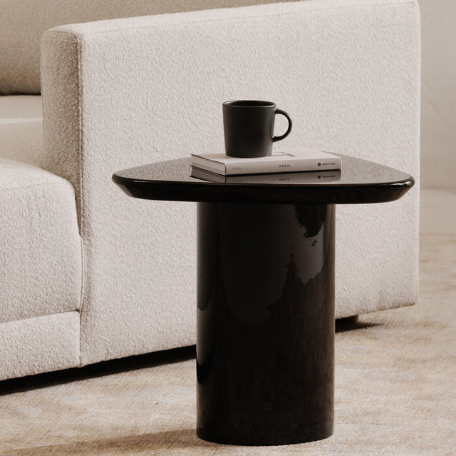 Table d’appoint moderne noire brillante avec un plateau organique et une base cylindrique, présentée à côté d’un canapé clair dans un intérieur minimaliste.