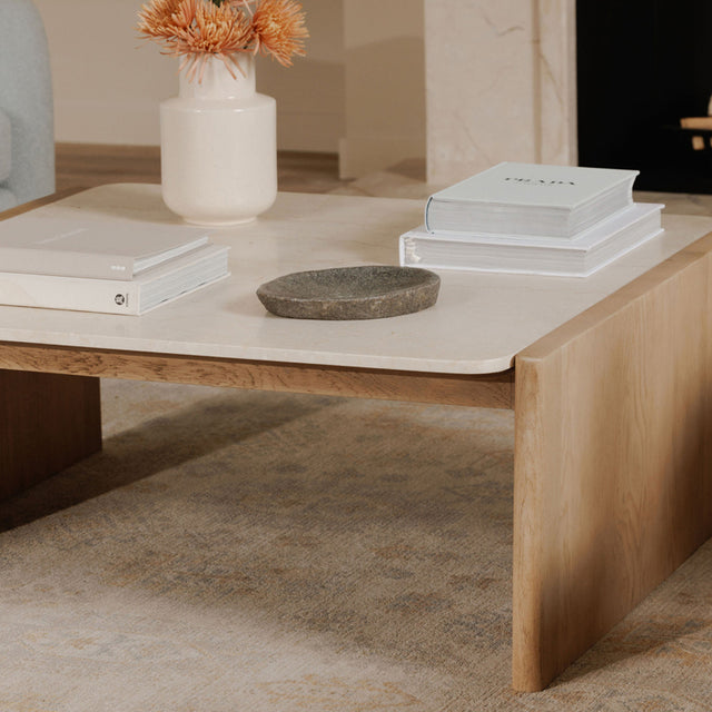 Table basse moderne en bois clair avec plateau beige effet pierre, décoree de livres et d’un vase.