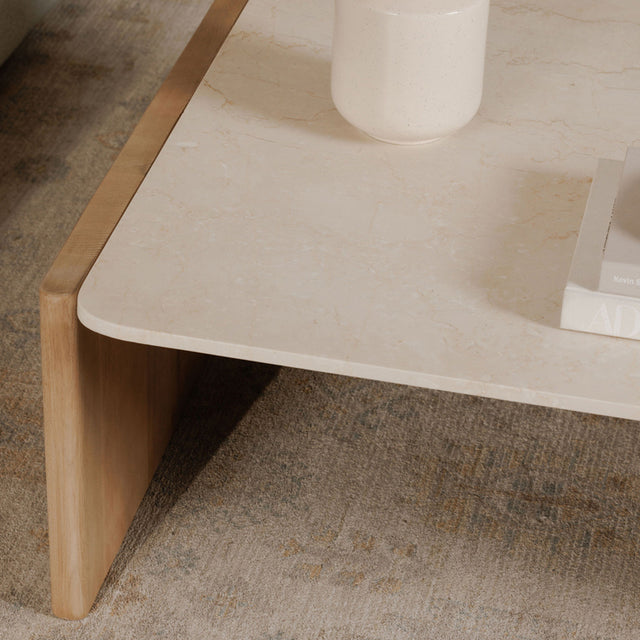 Détail d’un coin de table basse avec plateau beige effet pierre et structure en bois clair.