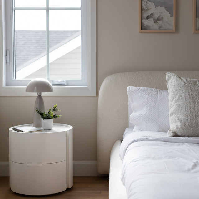 chambre minimaliste avec table de nuit blanche, lampe moderne et plante verte près d'un lit aux draps blancs.