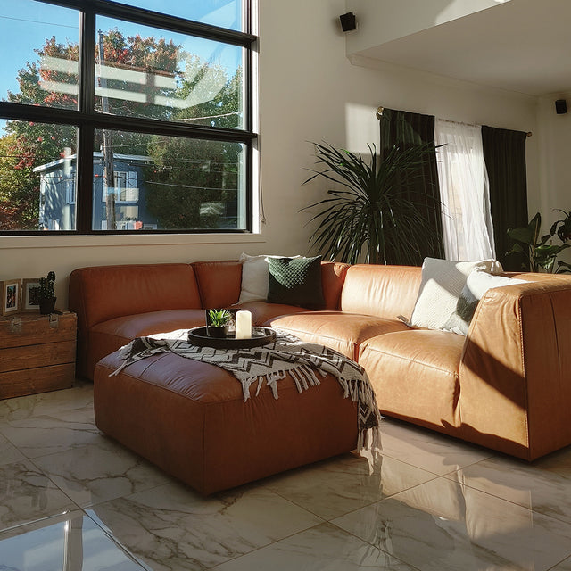 salon moderne avec canape d’angle en cuir brun, tapis clair, deux cadres muraux et plante verte dans un pot blanc.