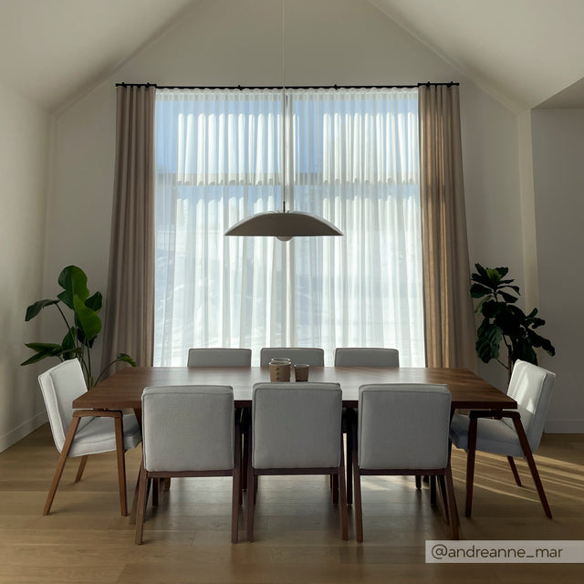 Salle a manger moderne avec grande table en bois, chaises grises et luminaire suspendu devant large fenetre.