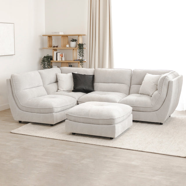 Canapé sectionnel gris clair avec ottomane assortie, coussins noirs et blancs, sur un grand tapis beige dans un salon lumineux et minimaliste.