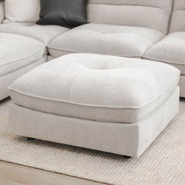 Ottomane grise moderne assortie au canapé sectionnel, posée sur un grand tapis beige texturé.