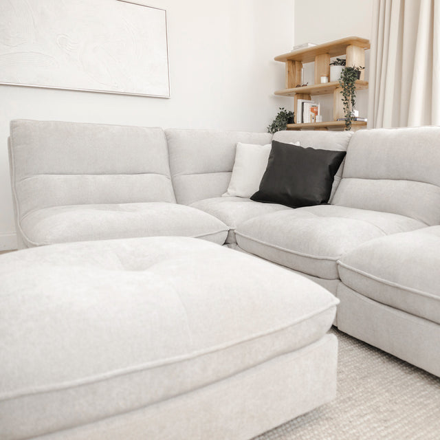 Canapé sectionnel gris clair moderne avec ottomane assortie et coussins décoratifs noirs et blancs, dans un salon lumineux au style minimaliste.