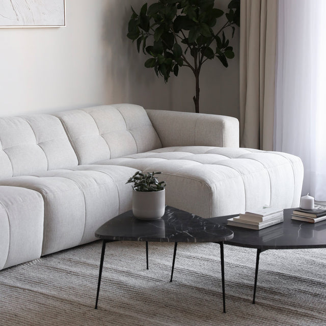 Canapé d’angle beige clair au design moderne et matelassé, accompagné de tables basses noires en marbre et d’un tapis texturé gris clair.