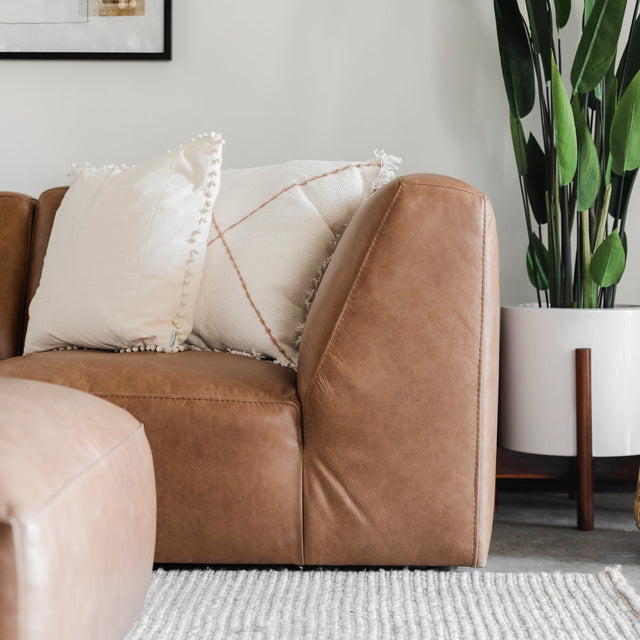 gros plan sur un canapé en cuir brun avec accoudoir incliné, coussins décoratifs clairs et une plante verte en pot à côté.