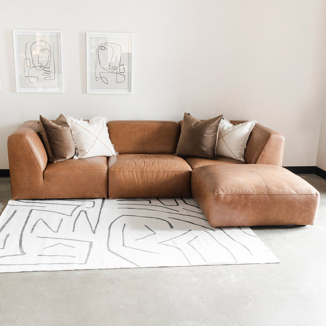 canapé d’angle en cuir brun clair avec des coussins crème et taupe, disposé sur un tapis blanc à motifs graphiques gris, devant un mur blanc orné de deux cadres minimalistes aux lignes abstraites.