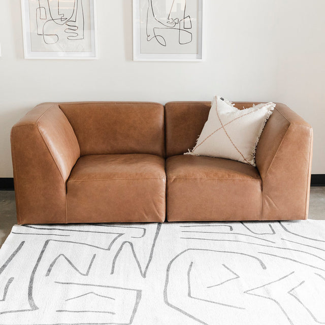 canapé deux places en cuir brun avec coussin décoratif clair, posé sur un tapis blanc à motifs abstraits gris, et deux cadres minimalistes accrochés au mur.