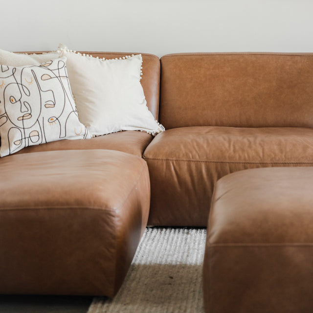 canapé d’angle en cuir brun avec assises larges, coussins décoratifs beiges et tapis clair à texture épaisse.
