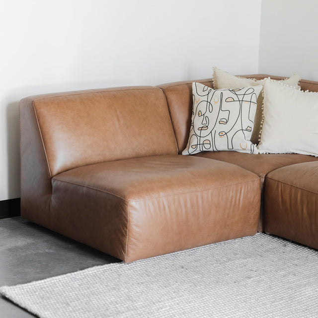 canapé d’angle en cuir brun avec assises larges, coussins décoratifs beiges et tapis clair à texture épaisse.