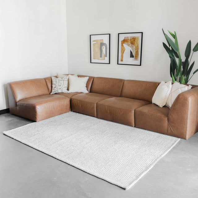 salon moderne avec canapé d’angle en cuir brun, tapis clair, deux cadres muraux et plante verte dans un pot blanc.