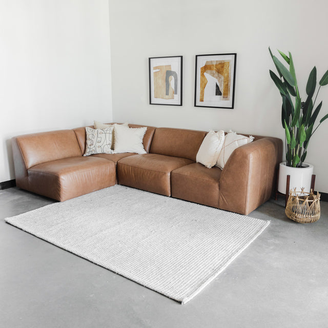 canapé d’angle en cuir brun avec plusieurs coussins beiges et bruns, disposé sur un tapis blanc à motifs abstraits gris, devant un mur clair décoré de deux cadres minimalistes