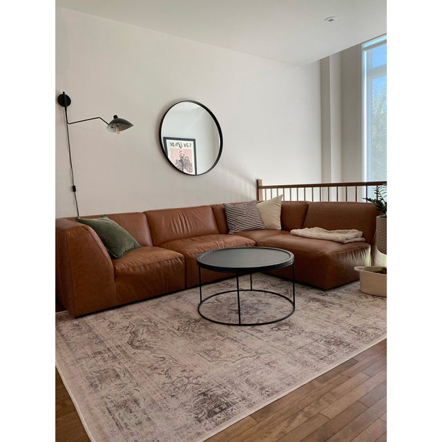 salon moderne avec canapé d’angle en cuir brun, table basse ronde noire, grand miroir mural circulaire et tapis clair à motifs discrets.