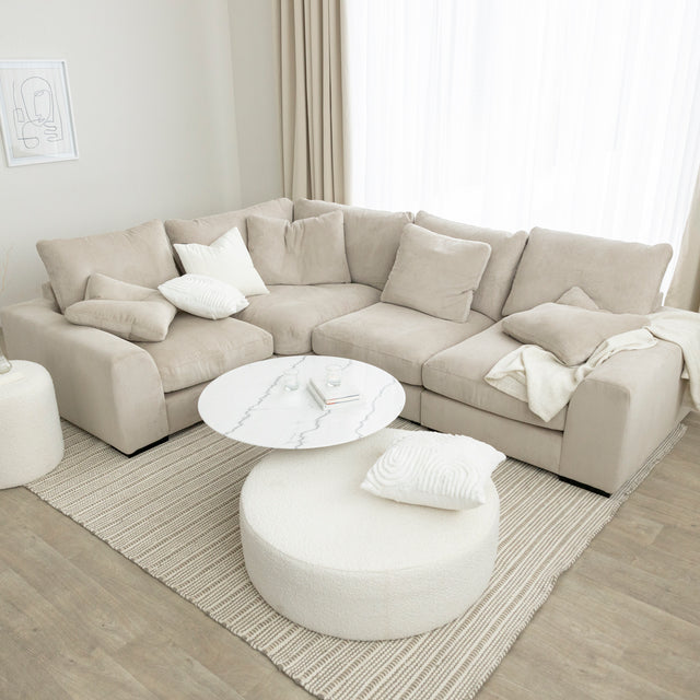 Canape d angle beige moelleux avec table basse blanche et pouf rond en bouclette sur tapis clair.