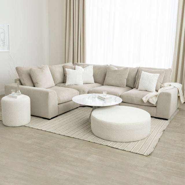 Salon lumineux avec canapé d’angle beige moelleux, table basse ronde blanche et pouf en bouclette sur tapis à rayures.