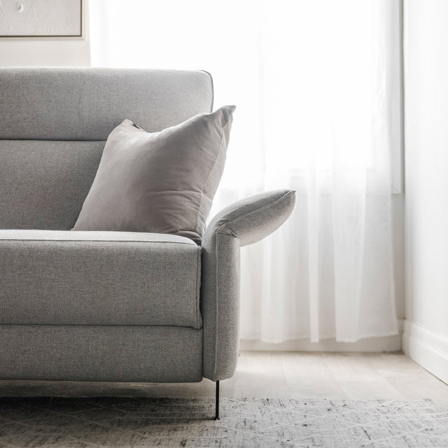 vue detaillee du canape gris clair avec accoudoir releve et coussin assorti devant une fenetre lumineuse.